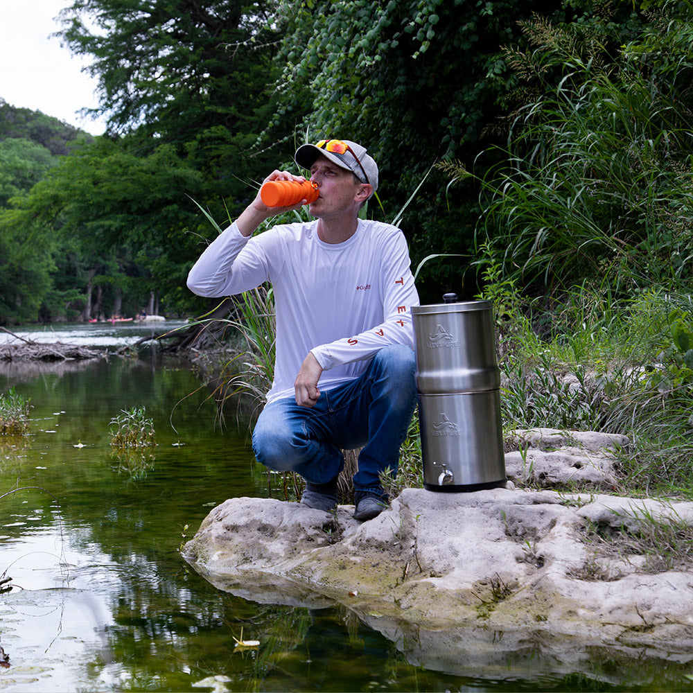 Man drinking from a collapsible sports bottle kneeling down next to the Alexapure Pro. A green water stream flows in the foreground.