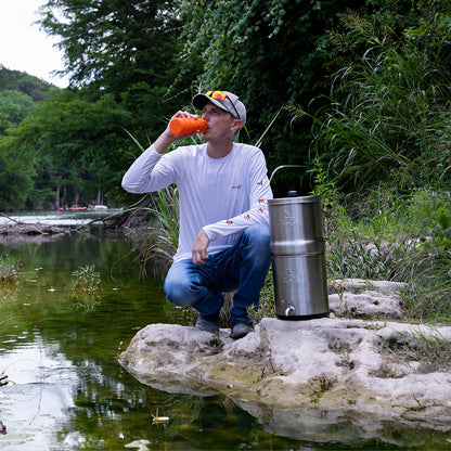 Man drinking from a collapsible sports bottle kneeling down next to the Alexapure Pro. A green water stream flows in the foreground. 